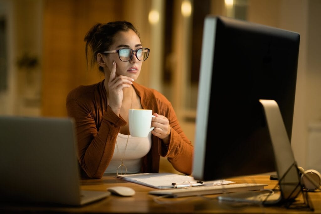Young pensive woman drinking tea while reading e-mail on computer at home.